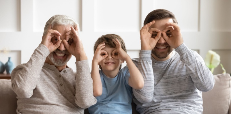 A smiling boy, with his dad and grandfather, on a sofa at home having fun making eyeglasses with their hands around their eyes