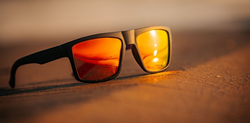 A pair of sunglasses resting on the sand reflects a glowing orange sunset