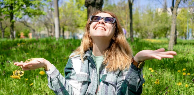 A young woman sat outside in the grass wearing sunglasses, looking up towards the sky
