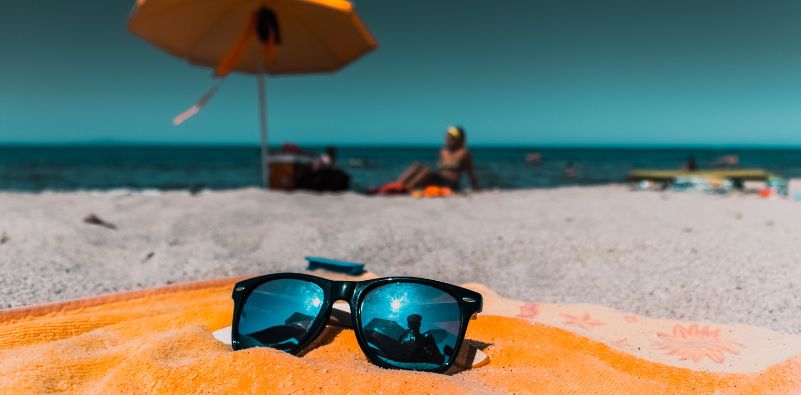 Black sunglasses resting on an orange beach towel on a sunny sandy beach, with the ocean, beach umbrella, and people relaxing in the background