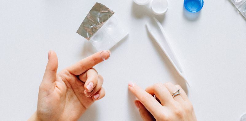 A flat lay of a pair of contact lenses and case, featuring a pair of hands with a contact lens on the fingertip of the left hand.