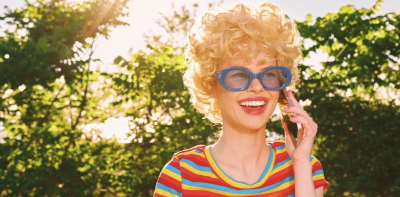 A woman wearing bold blue oval statement glasses and a colourful striped top smiling outdoors.