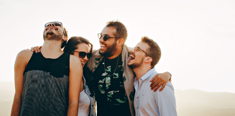 A group of friends hugging and laughing, wearing eyeglasses and sunglasses outdoors
