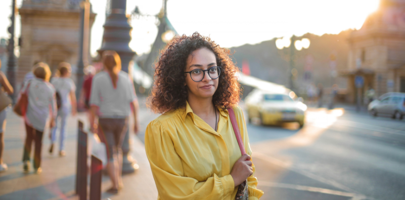 A young woman walking outside in the street, wearing eyeglasses and a yellow shirt
