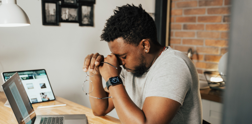 A man working at his laptop in a home environment, holding his eyeglasses in one hand and resting his head against the other