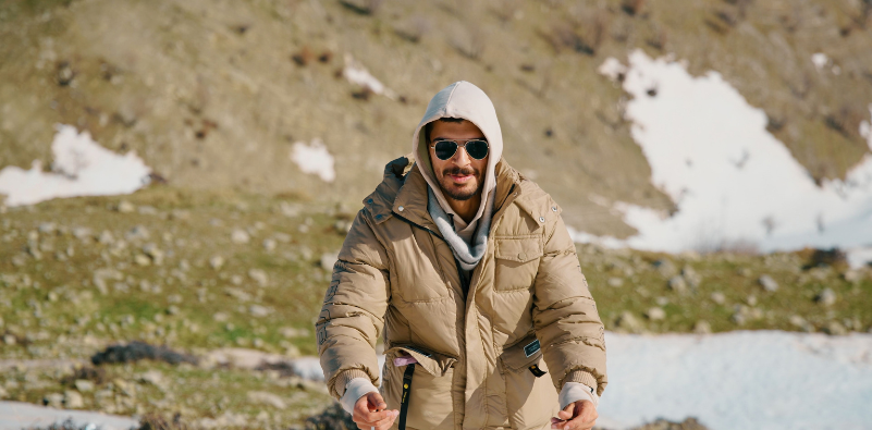 A man wearing a coat, hoodie and sunglasses, walking in the mountains with snow in the background